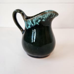May include: A small, dark green ceramic pitcher with a handle and a spout. The rim of the pitcher has a turquoise glaze. The pitcher is glossy and reflects light. The overall shape is rounded.