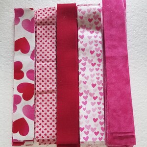 May include: A collection of fabric strips in shades of pink and red. The strips feature various patterns, including large hearts, small hearts, and solid colors. Ideal for crafting projects, the fabrics are arranged side by side.
