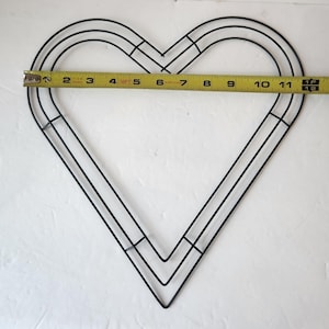 Puede incluir: Marco de corona en forma de corazón de metal negro con una cinta métrica amarilla en el centro. El marco mide aproximadamente 30 cm de ancho. El marco está hecho de finas varillas de metal y está diseñado para manualidades.