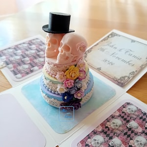 May include: A three-tier wedding cake decorated with pastel-coloured flowers and lace. The top tier features two pink sugar skulls, one wearing a black top hat. The cake is surrounded by a white background with the text "John & Carmia 7th November 2018".