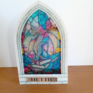 May include: A decorative arched frame featuring a stained glass style illustration of a dragon perched on a stack of books. The frame is light gray with a stone-like texture. A rectangular plaque at the base displays the name HETTIE in black text.