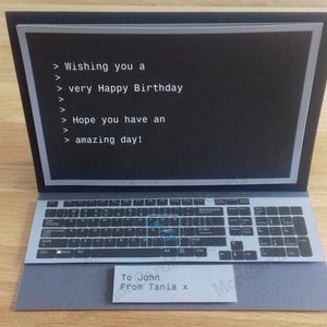 May include: A pop-up card shaped like a laptop computer with a black screen that says "Wishing you a very Happy Birthday Hope you have an amazing day!" The keyboard has a blue "Happy Birthday" message on the "Enter" key. The card is signed "To John From Tania x".