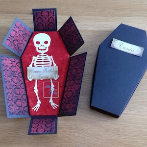 May include: A red velvet coffin box with a skeleton holding a sign that says "Happy Birthday Tania". The box is open and the skeleton is visible inside. The coffin box is decorated with a black and red damask pattern. A black coffin with a white label that says "Tania" is also in the image.