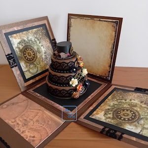 May include: A brown and black steampunk-themed box with a 3-tiered cake inside. The cake is decorated with black lace, gears, and flowers. The box is open and has a brown background with a map design.