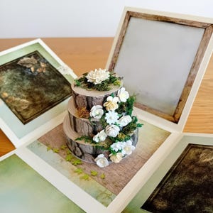 May include: A three-tiered wedding cake made to look like tree bark with white flowers and green leaves. The cake is in a white box with a brown frame.
