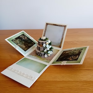 May include: A white and green paper box with a 3-tiered cake made of wood and white flowers. The box is decorated with lace and has a small window on the top.
