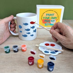 May include: A white mug with blue fish illustrations is being painted with a red fish using a paintbrush. The kit includes paint, a palette, and a box that reads "Paint Your Own Mug Kit."