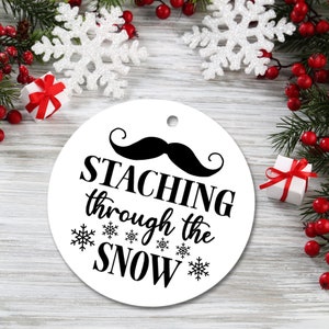 May include: White round Christmas ornament with black text that reads "STACHING through the SNOW" and a black mustache above the text.  The ornament is surrounded by snowflakes and red berries.