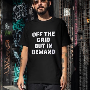 May include: A black short-sleeved t-shirt featuring the white text "OFF THE GRID BUT IN DEMAND" printed in a bold, stencil-style font across the chest. The garment has a crew neckline and a standard fit, providing a clean look for casual wear.