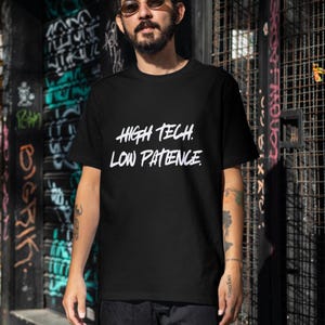 May include: A black short-sleeved t-shirt featuring the white text "HIGH TECH. LOW PATIENCE." printed in a bold, stylized font across the chest. The garment has a classic crew neckline and a straight hem, providing a simple and clean design.