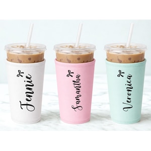 May include: Three iced coffee drinks in clear plastic cups with straws, each in a different colored sleeve. The sleeves are white, pink, and mint green, with the names "Jennie", "Samantha", and "Veronica" printed in black script.