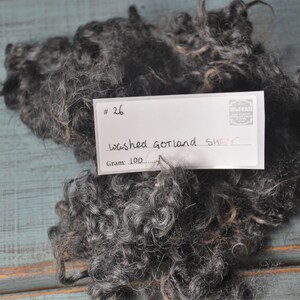 May include: A pile of dark gray washed Gotland sheep wool. The wool is curly and textured, with a mix of gray and light brown fibers. A white label with the text "#26 washed Gotland sheep" is on top of the wool.