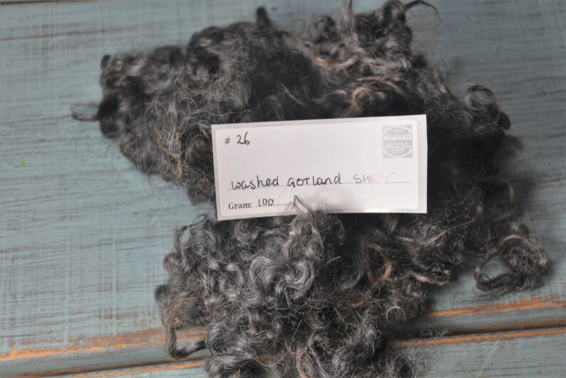 May include: A pile of dark gray washed Gotland sheep wool. The wool is curly and textured, with a mix of gray and light brown fibers. A white label with the text "#26 washed Gotland sheep" is on top of the wool.