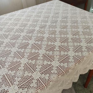 Puede incluir: Un mantel de ganchillo de color crema con un patrón geométrico detallado. El mantel es rectangular y cubre una mesa de madera. Los bordes tienen un ribete festoneado. El intrincado diseño añade un toque de elegancia a la mesa.