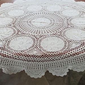 Op de afbeelding: Een rond, gebroken wit gehaakt tafelkleed met een centraal cirkelvormig motief omringd door een ring van acht kleinere, stervormige bloemmotieven. Het ontwerp bevat een ingewikkelde opengewerkte rand met een decoratieve schulprand.