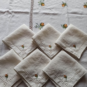May include: Set of six white linen napkins with embroidered orange and green flowers. The napkins have a scalloped edge and are folded in a square shape.