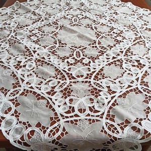 May include: A white lace tablecloth featuring an intricate cutwork design with repeating floral motifs and interconnected scrollwork patterns. The fabric displays a delicate openwork structure with embroidered leaf details throughout the entire piece.