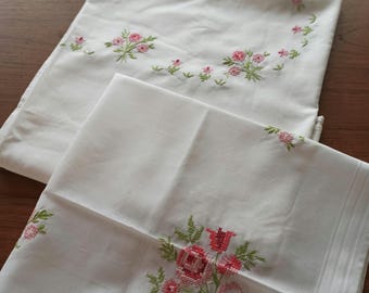 Op de afbeelding: Twee witte geborduurde tafelkleden met roze en groene bloemenmotieven. Een tafelkleed heeft een cirkelvormig ontwerp met kleine bloemen, terwijl de andere een groter boeket bloemen in de hoek heeft.