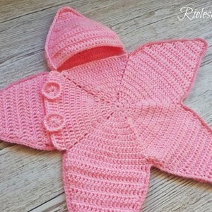 May include: A handmade, pink crochet baby outfit, shaped like a starfish, with a matching hat. The outfit has button closures and is displayed on a wooden surface. The item is a baby garment.