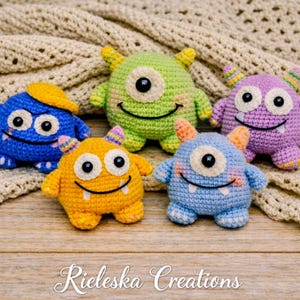 May include: A collection of five colorful, crocheted monster toys. Each monster has a unique color, including blue, green, yellow, and purple, with black eyes and smiling mouths. The toys are displayed on a wooden surface with a crocheted blanket in the background.