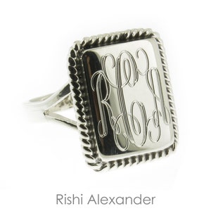 May include: A silver ring with a square-shaped band featuring a rope-like border. The band has a monogrammed inscription with the letters "RLA".