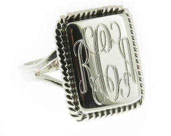 May include: A silver ring with a square-shaped band featuring a rope-like border. The band has a monogrammed inscription with the letters "RLA".