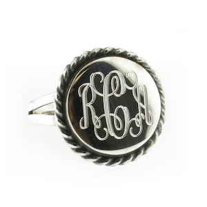 May include: A silver ring with a rope-like design around the edge of the band. The ring has a circular face with a monogram engraved in the center.
