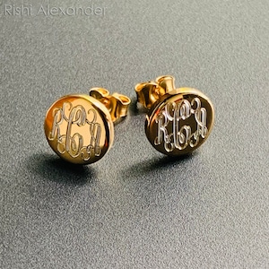 May include: A pair of gold-colored stud earrings. Each earring is round with a monogram design in an elegant script. The earrings are displayed on a gray surface.