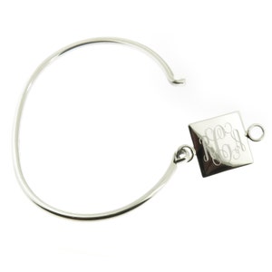 May include: A silver-colored bangle bracelet with a square charm. The charm is engraved with a monogram in a cursive font. The bracelet has an open design with a hook closure. The jewelry is displayed on a white background.