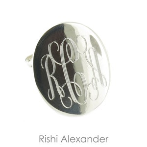 May include: A silver oval cufflink with a monogram of the letters "RJA" engraved in a script font.