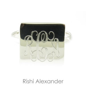 May include: A silver ring with a square-shaped band featuring a monogrammed design. The monogram is engraved in a script font.