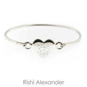 May include: A silver heart-shaped bangle bracelet with a personalized monogram. The bracelet is made of a thin, delicate metal and has a simple, elegant design.