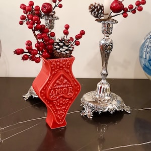 Könnte beinhalten: Zwei silberne Kerzenleuchter mit weißen Kerzen und roten Beerenzweigen. Eine rote Keramikvase mit der Aufschrift "AMOR" steht in der Mitte des Bildes. Die Vase ist mit roten Beeren und einer Kiefernzapfen gefüllt.