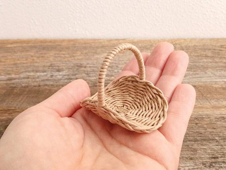 Może przedstawiać: Miniaturowy pleciony koszyk w kolorze jasnego beżu z pojedynczym łukowatym uchwytem. Koszyk posiada okrągłą podstawę i pleciony brzeg, wykonany z cienkich, elastycznych pask&oacute;w materiału w ciasny, sp&oacute;jny wz&oacute;r, przeznaczony do użytku w małej skali.
