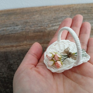 Może przedstawiać: Miniaturowy biały pleciony koszyk z pojedynczym łukowatym uchwytem, zawierający mały bukiet czterech jasnor&oacute;żowych sztucznych pąk&oacute;w r&oacute;ż związanych cienką, p&oacute;łprzezroczystą brązową wstążką, prezentowany jako delikatny przedmiot dekoracyjny.