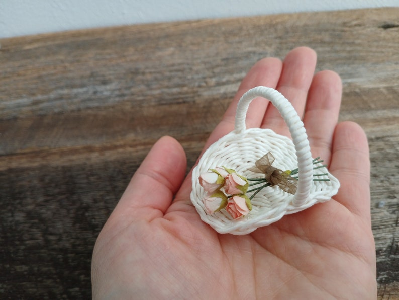 Może przedstawiać: Miniaturowy biały pleciony koszyk z pojedynczym łukowatym uchwytem, zawierający mały bukiet czterech jasnor&oacute;żowych sztucznych pąk&oacute;w r&oacute;ż związanych cienką, p&oacute;łprzezroczystą brązową wstążką, prezentowany jako delikatny przedmiot dekoracyjny.
