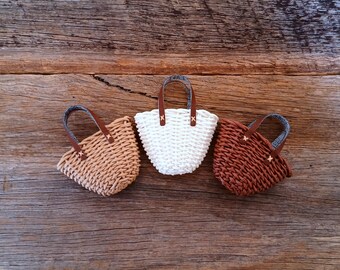 May include: Three small woven tote bags in tan, white, and brown, each with a brown leather handle and decorative stitching. The bags are arranged on a rustic wooden surface, showcasing their handmade quality.