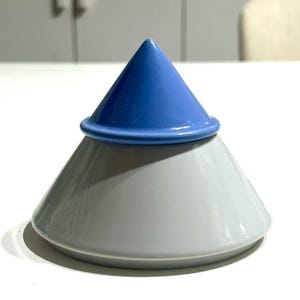 Op de afbeelding: Een tweedelig keramisch object met een brede, lichtgrijze conische basis en een kleinere, gladde blauwe conische deksel die er bovenop rust. Het object heeft een glanzende afwerking, een geometrische vorm en een strak, gestapeld ontwerp.