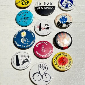 Może przedstawiać: Kolekcja dwunastu okrągłych przypinek z różnymi grafikami politycznymi i społecznymi, zawierająca napisy takie jak Greenpeace, ik fiets stil & schoon, Amnesty International oraz Atoomenergie? Nee bedankt, w różnych kolorach.