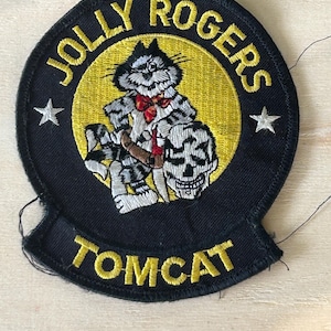 Puede incluir: Un parche bordado circular con borde negro y centro amarillo. Presenta un gato de dibujos animados con pajarita roja y cinturón marrón, de pie junto a una calavera. El texto dice JOLLY ROGERS en la parte superior y TOMCAT en la inferior, con dos estrellas blancas.