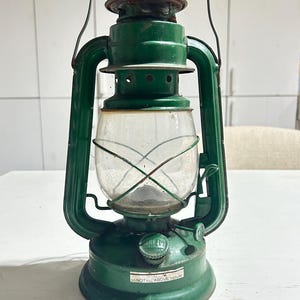 May include: A green metal oil lantern featuring a clear glass globe protected by a wire cage. The base includes a fuel cap and a white label with the text DO NOT FILL ABOVE THIS LINE. A wire handle is attached to the top for carrying purposes.