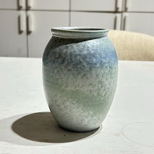 Può includere: Un piccolo vaso in ceramica con corpo arrotondato e apertura stretta. L'esterno presenta uno smalto maculato che sfuma da un blu intenso sul bordo superiore a una finitura verde salvia tenue e bianca verso la base, creando un effetto sfumato.
