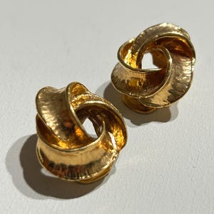 Op de afbeelding: Een paar goudkleurige oorbellen met een sculpturaal, geknoopt ontwerp en een getextureerd, gehamerd oppervlak. De oorbellen hebben een ronde, wervelende vorm met een open midden, waardoor het licht reflecteert op de onregelmatige metalen afwerking.