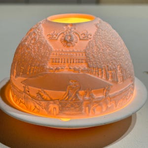 May include: A white porcelain dome-shaped tea light candle holder featuring a relief carving of a classical building, trees, and a horse-drawn chariot. The translucent material glows with a warm orange light from within, resting on a circular base.