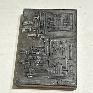 以下が含まれることがあります： 機械の複雑な技術図面が描かれた長方形の金属製活版印刷用プレート。銀色の表面には、精巧な彫刻線、機械部品、およびデザイン全体にわたるさまざまな数値ラベルが表示されています。