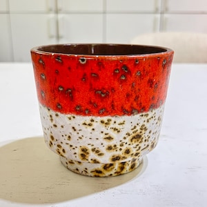 May include: A ceramic planter featuring a vibrant orange upper section with dark speckles and a white lower section with brown mottled spots. The vessel has a cylindrical shape with a slightly tapered base and a smooth, dark brown interior rim.