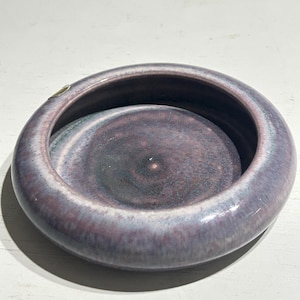 Könnte beinhalten: Eine runde Keramikschale mit einem dicken, geschwungenen Rand. Die Glasur weist eine gesprenkelte Mischung aus tiefen Violett-, staubigen Lavendel- und dunklen Anthrazittönen auf, die eine glatte, glänzende Oberfläche mit subtilen Farbvariationen erzeugt.