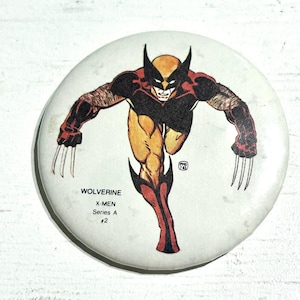 Peut inclure: Un badge rond illustrant le personnage Wolverine dans un costume jaune, noir et marron avec des griffes déployées. Le texte indique WOLVERINE X-MEN Series A #2, accompagné d'un petit tampon PMS 83 visible sur le côté droit de l'image.