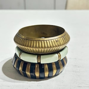 Pode incluir: Uma pilha de três braceletes. O bracelete superior é uma faixa larga de latão texturizado. O bracelete central apresenta segmentos retangulares lisos de cor verde menta pálido separados por pequenos espaçadores de latão.