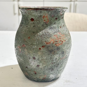 Può includere: Un vaso in ceramica con una superficie ruvida e maculata nei toni del verde salvia, grigio antracite e rosso ruggine. Il recipiente presenta un corpo ampio e arrotondato che si restringe verso la base e si allarga sul bordo superiore, con finitura rustica.